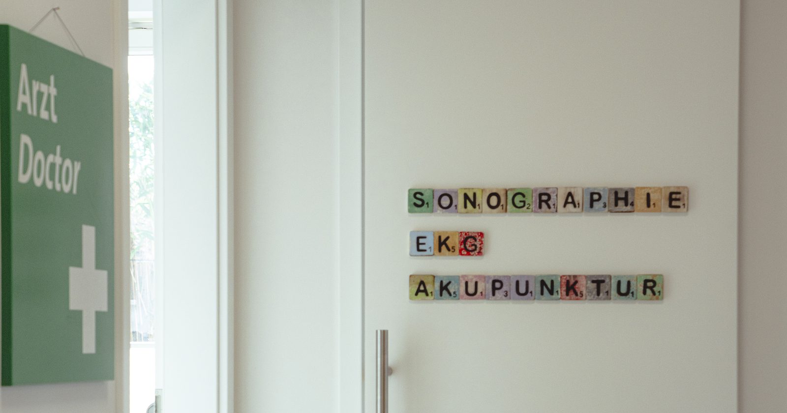 Praxisflur mit dem Hinweisschild Arzt Doctor, an der Tür bunte Buchstaben mit den Worten Sonographie, EKG und Akupunktur